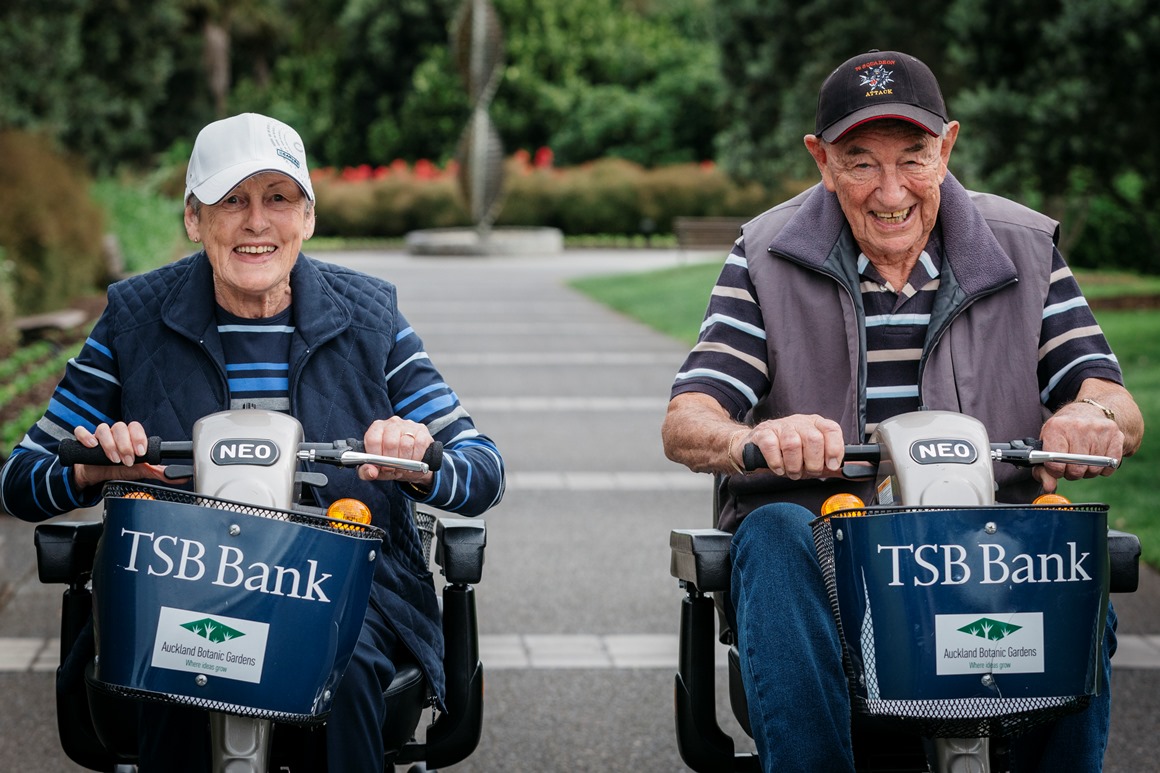 An older woman and man smile as they drive two mobility scooters side by side up a path at Auckland Botanic Gardens