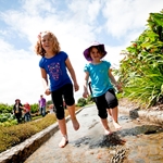 Taken from low down the photo shows two children running barefoot through a channel of water on a warm sunny day at Auckland Botanic Gardens