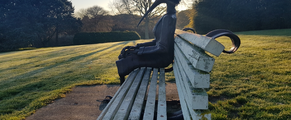 It is dawn in winter in the gardens and a soft sun is rising over the horizon silhouetting a scultpure of a bird woman. She is sitting on a park bench looking north with her hands clasped, perhaps a little lonely, but also at home.