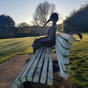 It is dawn in winter in the gardens and a soft sun is rising over the horizon silhouetting a scultpure of a bird woman. She is sitting on a park bench looking north with her hands clasped, perhaps a little lonely, but also at home.