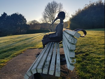 It is dawn in winter in the gardens and a soft sun is rising over the horizon silhouetting a scultpure of a bird woman. She is sitting on a park bench looking north with her hands clasped, perhaps a little lonely, but also at home.