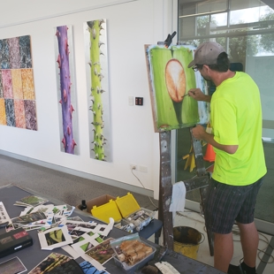 Artist Neal Palmer is working at his easel. He has his back to the camera and paintbrush in hand. He is applying paint to a work in progress that will be a close up of a thorn. In the background are some of Neal's finished works of bark rubbings and large scale plant stems covered in thorns. In the foreground are an apparently chaotic arrangement of papers and photographs Neal has taken of various plants during his time as artist in residence at Auckland Botanic Gardens.